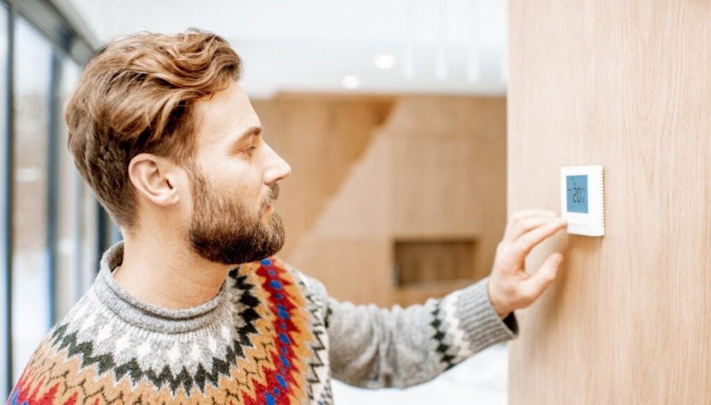 Man in sweater feeling cold adjusting room temperature with electronic thermostat at home